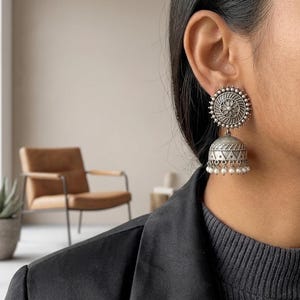 May include: A silver-toned jhumka earring featuring a circular stud with a central floral motif and a bell-shaped drop pendant. The lower edge of the bell is decorated with a row of small, white, spherical beads hanging from the base.