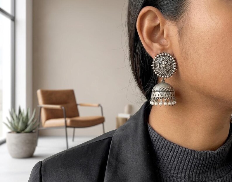 May include: A silver-toned jhumka earring featuring a circular stud with a central floral motif and a bell-shaped drop pendant. The lower edge of the bell is decorated with a row of small, white, spherical beads hanging from the base.