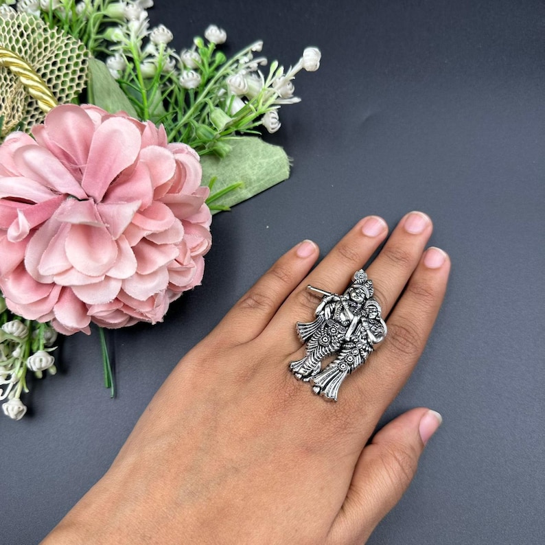 Puede incluir: Un anillo plateado que representa dos figuras, posiblemente deidades, con ropa y ornamentaci&oacute;n detalladas. El anillo se lleva en una mano, sobre un fondo oscuro, con una flor rosa y vegetaci&oacute;n en la esquina superior izquierda.