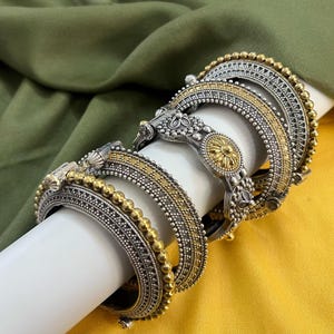 Könnte beinhalten: Mehrere silber- und goldfarbene Armreifen mit aufwendigen Designs werden auf einem weißen zylindrischen Objekt präsentiert. Die Armreifen weisen eine Mischung aus Texturen auf, darunter Perlen- und gemusterte Oberflächen. Der Hintergrund besteht aus olivgrünem und gelbem Stoff.