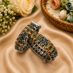 Puede incluir: Un par de brazaletes articulados con un acabado bicolor en plata y oro. El trabajo en metal presenta un intrincado patrón de celosía acentuado con pequeñas piedras preciosas redondas de color verde oscuro incrustadas en el diseño.