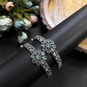 Può includere: Due braccialetti d'argento con un design floreale. Ogni braccialetto presenta piccole pietre preziose blu chiaro disposte a forma di fiore. I braccialetti sono esposti su un oggetto cilindrico nero, con fiori sullo sfondo.