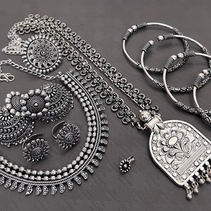 Pode incluir: Um conjunto de joias prateadas que inclui um colar com um pingente, brincos, um anel e quatro pulseiras. O colar tem um pingente grande e adornado com um design floral. Os brincos são grandes e redondos com detalhes intrincados. O anel é uma banda simples com uma pequena pedra redonda. As pulseiras são de diferentes tamanhos e têm uma variedade de designs.