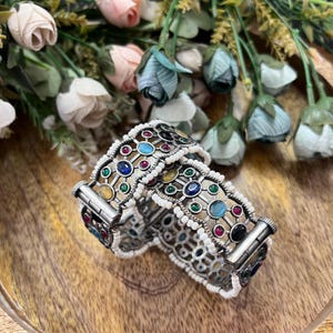 以下が含まれることがあります： カラフルな宝石と白いビーズのアクセントが付いた、2つのシルバーカラーのバングル。バングルは、赤、青、緑、黄、黒の石を使った幾何学模様のデザインです。ジュエリーは木製の表面に展示されています。