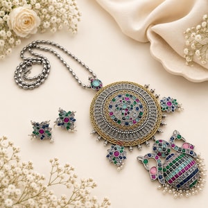 May include: A silver-toned necklace featuring a large circular pendant with intricate gold and silver filigree, inlaid with multicolored gemstones. The set includes matching stud earrings, each adorned with a central pink stone surrounded by blue and green accents, and a silver bead chain.