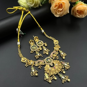 Può includere: Un set di collana e orecchini color oro con intricati disegni floreali e geometrici. La collana presenta un pendente centrale con accenti verdi e rossi, e gli orecchini hanno dettagli coordinati. I gioielli sono esposti su una superficie scura.