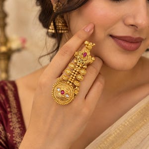 Op de afbeelding: Een goudkleurige statement ring met een langwerpig ontwerp, ingewikkeld filigraanwerk, kleine muntachtige accenten, een centraal rond medaillon met een rode steen en een witte parel, en een druppelvormige rode steen aan de bovenzijde.