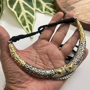 Puede incluir: Un collar gargantilla plateado y dorado con intrincados adornos y elementos decorativos. El collar tiene un cierre de cord&oacute;n negro. El dise&ntilde;o presenta una mezcla de tonos plateados y dorados, creando un contraste llamativo.