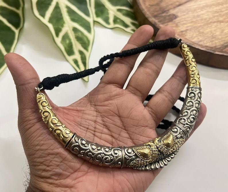 Puede incluir: Un collar gargantilla plateado y dorado con intrincados adornos y elementos decorativos. El collar tiene un cierre de cord&oacute;n negro. El dise&ntilde;o presenta una mezcla de tonos plateados y dorados, creando un contraste llamativo.