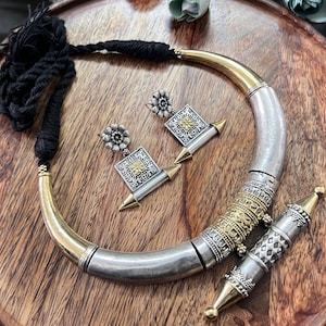 Puede incluir: Un conjunto de collar y pendientes plateados y dorados con un colgante a juego. El collar tiene un diseño grueso y curvado con un cordón negro. Los pendientes y el colgante presentan diseños cuadrados y florales. Las joyas se exhiben sobre una superficie de madera.