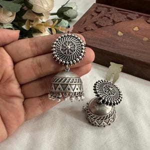 May include: A pair of silver earrings with a round, ornate design. The earrings have a dangling, bell-shaped charm with a geometric pattern.