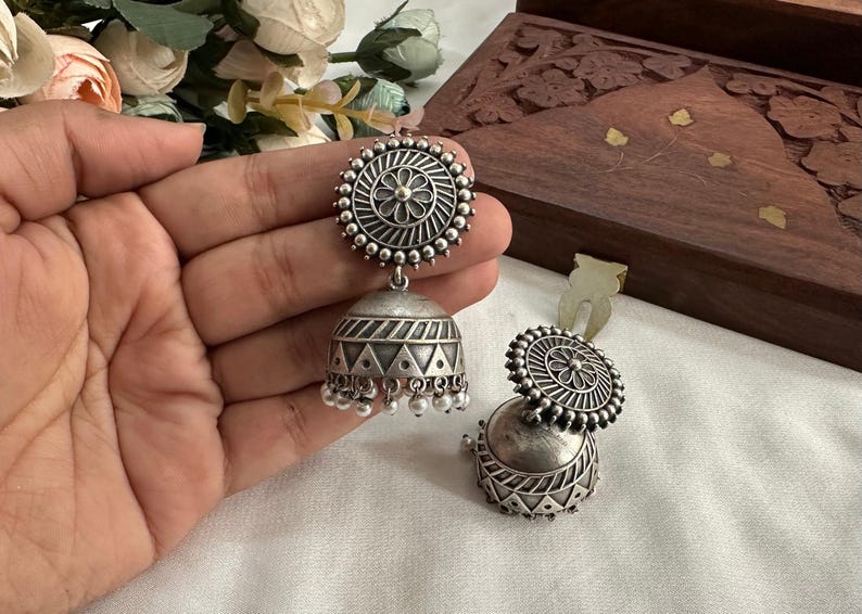 May include: A pair of silver earrings with a round, ornate design. The earrings have a dangling, bell-shaped charm with a geometric pattern.