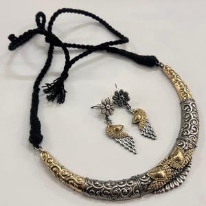 Puede incluir: Un conjunto de collar y pendientes plateado y dorado. El collar tiene un dise&ntilde;o curvo con patrones intrincados y un cord&oacute;n negro. Los pendientes presentan tachuelas florales y colgantes en forma de pluma. El conjunto de joyas se muestra sobre una superficie blanca.