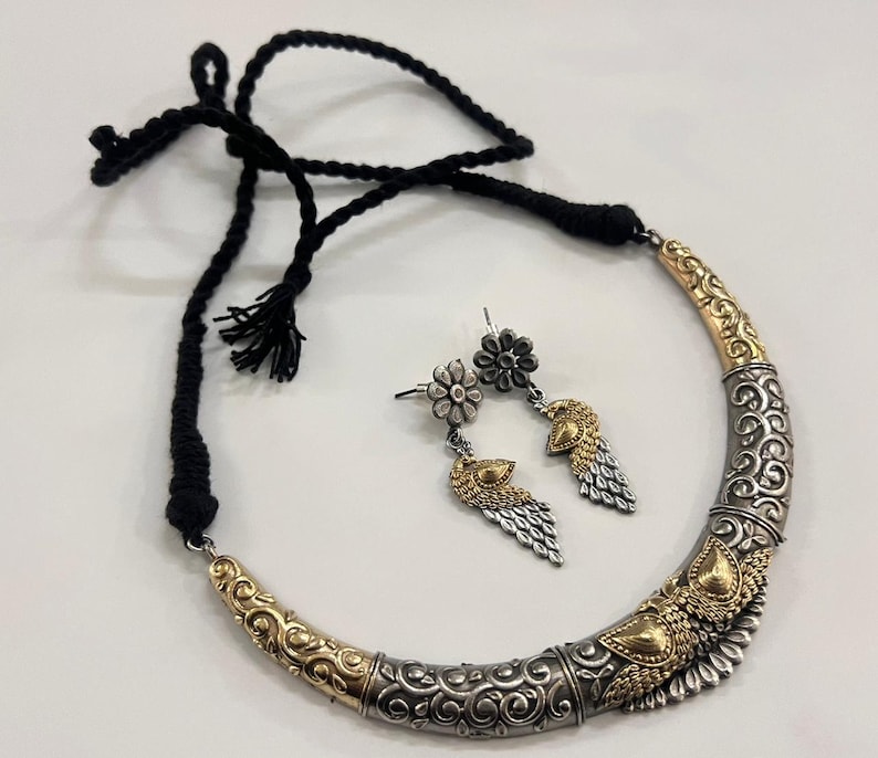 Puede incluir: Un conjunto de collar y pendientes plateado y dorado. El collar tiene un dise&ntilde;o curvo con patrones intrincados y un cord&oacute;n negro. Los pendientes presentan tachuelas florales y colgantes en forma de pluma. El conjunto de joyas se muestra sobre una superficie blanca.