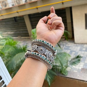 May include: A set of four silver bangles with green gemstone accents. The bangles are stacked on a person's wrist.