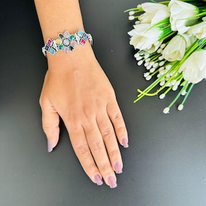 May include: Silver bracelet with a floral design, set with various coloured gemstones. The bracelet is on a hand with painted nails. The background is dark, with white flowers.