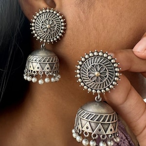 May include: Silver-toned earrings with a floral design and small pearl accents. The earrings feature a circular stud with a flower and a bell-shaped drop with geometric patterns. The earrings are a traditional style.