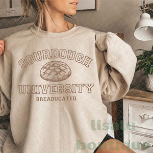 Puede incluir: Una sudadera beige con el texto "SOURDOUGH UNIVERSITY BREADUCATED" y un dibujo de una hogaza de pan de masa madre.