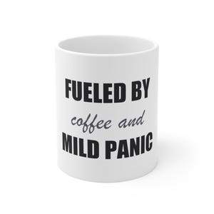 May include: A white ceramic mug featuring the black and grey text FUELED BY coffee and MILD PANIC printed on the front. The mug has a cylindrical shape with a smooth finish and a wide opening at the top, designed for holding hot or cold beverages.