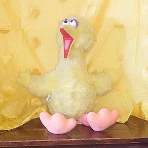 May include: A plush yellow bird with large blue eyes, an open red beak, and pink feet. The bird is sitting on a dark wooden surface against a yellow background. The bird's arms are outstretched.
