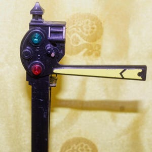 May include: A black and yellow model train signal with a green and red light. The signal arm is yellow with a black arrow. The signal has a small roof and is mounted on a black post.