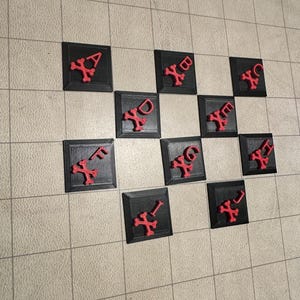 Puede incluir: Un conjunto de 10 fichas cuadradas negras con símbolos rojos de calavera y huesos cruzados. Cada ficha tiene una letra de la A a la J.