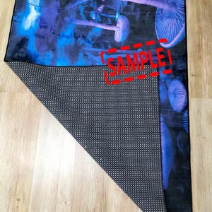 Puede incluir: Una alfombra de ba&ntilde;o azul y morada con un dise&ntilde;o de hongos. La alfombra tiene un respaldo antideslizante negro. La alfombra est&aacute; doblada por la mitad y la parte posterior es visible. La alfombra tiene la palabra "SAMPLE" impresa en ella.