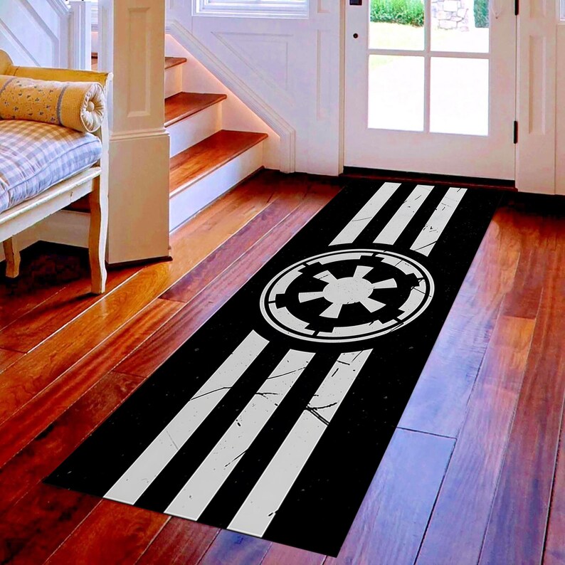 May include: Black and white Star Wars Imperial symbol area rug. The rug features a black background with three white stripes and the Imperial symbol in the center. The rug is rectangular and placed on a hardwood floor.