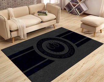 May include: A gray area rug with a blue Rebel Alliance symbol in the center. The symbol is surrounded by a circle of text in a dark blue color. The rug has two blue stripes running across it.