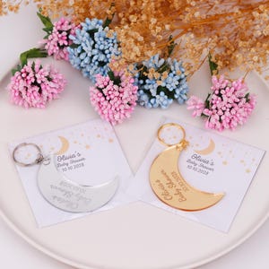 May include: Two crescent moon keychains, one silver and one gold, with the text "Olivia's Baby Shower 10.10.2028". The keychains are on a white plate with pink and blue floral decorations.
