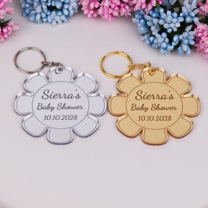 May include: Two flower-shaped keychains, one silver and one gold, with the text "Sierra's Baby Shower 10.10.2028" engraved on them. Each keychain has a metal ring for attaching keys. The background features colorful flowers.