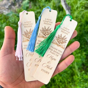 Può includere: Segnalibri in legno con nappe rosa, blu e verdi. Ogni segnalibro presenta il testo "A New Chapter Begins" e "Thank you for being a part of" seguito da un nome e "Story". I segnalibri hanno un design floreale e di libro.