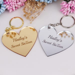 Op de afbeelding: Twee hartvormige sleutelhangers met een gespiegelde afwerking. De ene sleutelhanger is goud en de andere is zilver. Beide sleutelhangers hebben de tekst "Hailey's Sweet Sixteen" gegraveerd.