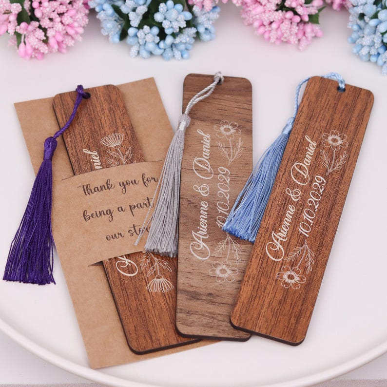 May include: Wooden bookmarks with floral designs and tassels in purple, gray, and blue. One bookmark is paired with a kraft paper card that reads "Thank you for being a part of our story." Two bookmarks are engraved with "Arienne & Daniel 10.10.2029".