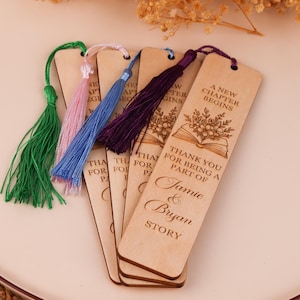 Pode incluir: Marcadores de livros de madeira com borlas coloridas. Os marcadores t&ecirc;m texto gravado que diz "A New Chapter Begins" e "Thank you for being a part of Jamie & Bryan Story". As borlas s&atilde;o verdes, rosa, azuis e roxas.