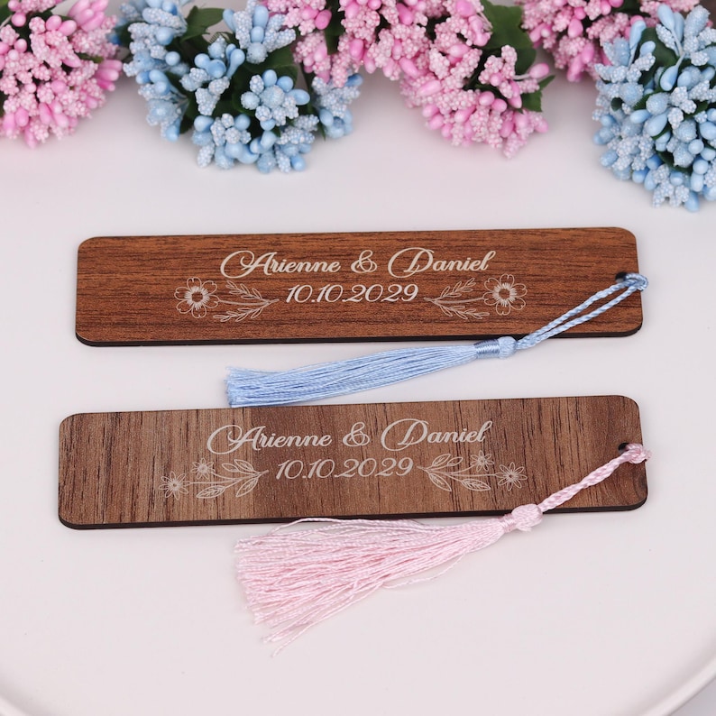 May include: Two wooden bookmarks with the names "Arienna & Daniel" and the date "10.10.2029" engraved in white. One bookmark has a light blue tassel, and the other has a pink tassel. The bookmarks are set against a white background with floral decorations.