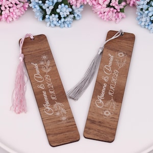 May include: Two wooden bookmarks with floral designs and the names "Arienna & Daniel" and the date "10.10.2029" printed in white. One bookmark has a pink tassel, and the other has a gray tassel. The bookmarks are rectangular with rounded corners.