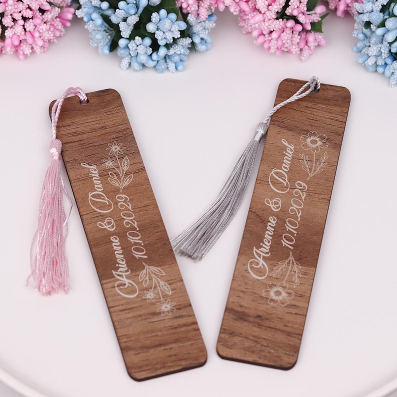 May include: Two wooden bookmarks with floral designs and the names "Arienna & Daniel" and the date "10.10.2029" printed in white. One bookmark has a pink tassel, and the other has a gray tassel. The bookmarks are rectangular with rounded corners.
