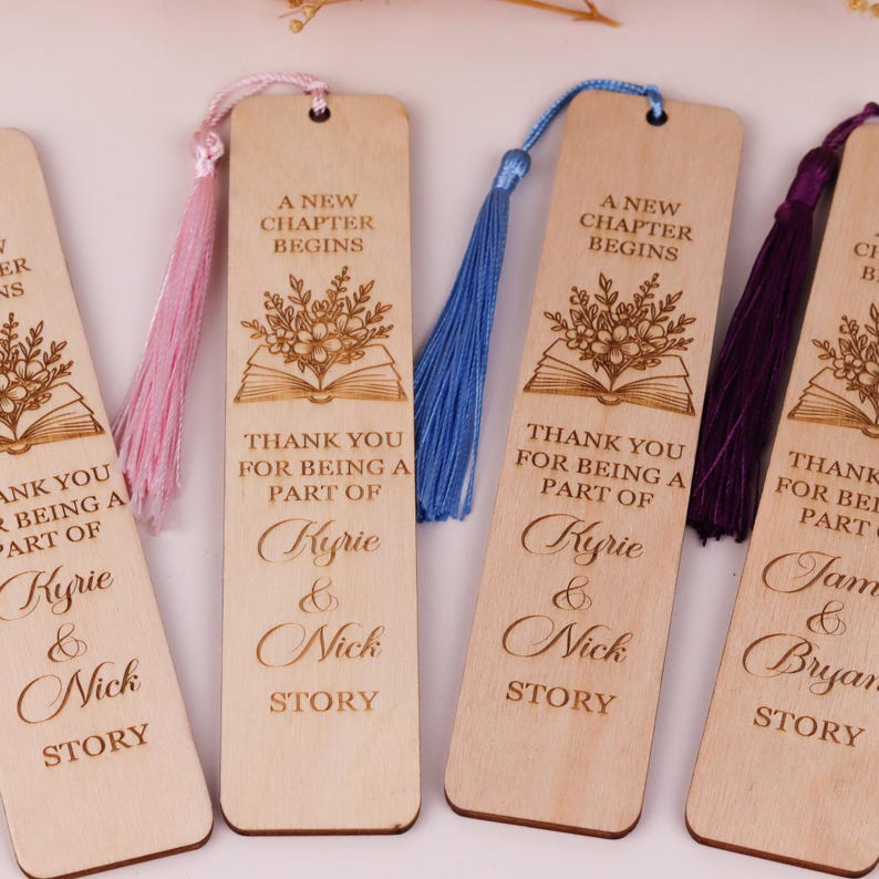 Pode incluir: Marcadores de livros de madeira com texto gravado e desenhos florais. Cada marcador tem uma borla de cor diferente. O texto diz "A New Chapter Begins" e "Thank you for being a part of Kyrie & Nick Story."