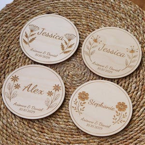 Può includere: Quattro sottobicchieri rotondi in legno con nomi incisi e disegni floreali. Ogni sottobicchiere presenta un nome diverso: Jessica, Alex e Stephanie. I nomi sono circondati da delicati motivi floreali e dalla data "10.10.2029".