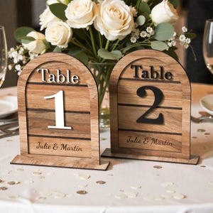 May include: Two wooden table number signs with arched tops. The signs feature the word "Table" above the numbers 1 and 2. The names "Julie & Martin" are printed below the numbers. The signs are on a white tablecloth with a floral arrangement in the background.