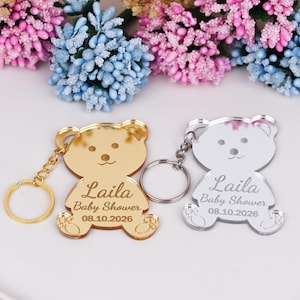 Op de afbeelding: Twee sleutelhangers in de vorm van een beer, één goudkleurig en één zilverkleurig, met de naam "Laila" en "Baby Shower 08.10.2026" gegraveerd. Elke sleutelhanger heeft een ring en een ketting. De achtergrond toont roze en blauwe bloemen.