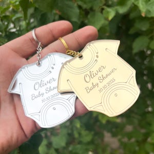 May include: Two silver and gold baby onesie shaped keychains with the name Oliver and the text "Baby Shower 10.10.2025".
