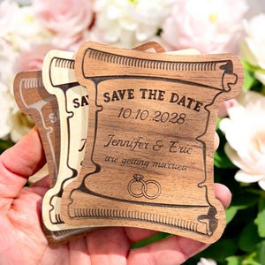 Könnte beinhalten: Holz-Save-the-Date-Ankündigung in Form einer Schriftrolle. Die Schriftrolle ist mit dem Text "Save the Date", "10.10.2028", "Jennifer & Eric are getting married" und zwei ineinander verschlungenen Ringen graviert.