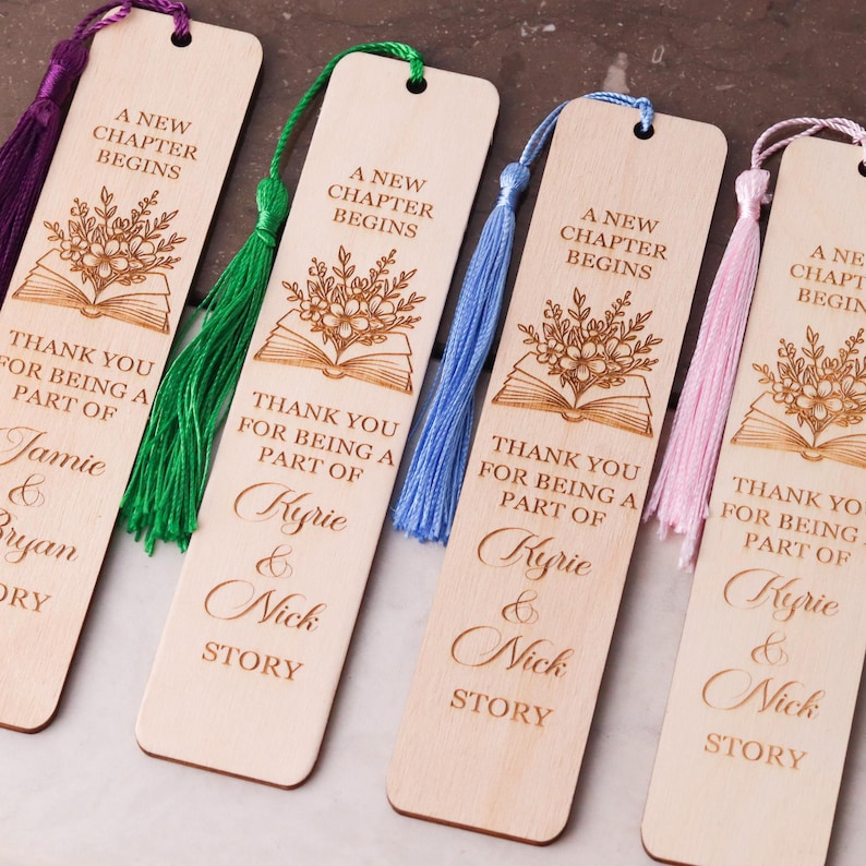 Pode incluir: Marcadores de livros de madeira com desenhos florais e de livros gravados, com o texto "A New Chapter Begins" e nomes personalizados. Cada marcador tem uma borla colorida, com op&ccedil;&otilde;es roxas, verdes, azuis e rosas.