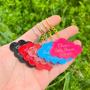 May include: A collection of cloud-shaped keychains in black, red, blue, and pink. Each keychain has a gold or silver ring and chain. The pink keychain reads "Oliver's Baby Shower 10.10.2029".
