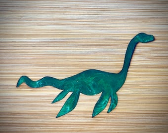 Puede incluir: Una silueta verde de una criatura marina mítica, el monstruo del lago Ness, con un cuello largo y aletas, sobre un fondo de madera.