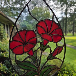 May include: A teardrop-shaped stained glass suncatcher featuring two vibrant red flowers with green leaves and stems. The piece is constructed with dark metal soldering lines and clear, textured glass panels that create a raindrop effect.