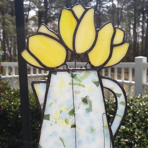 May include: A stained glass window hanging features a blue and white pitcher with a yellow tulip bouquet. The pitcher has a speckled design.