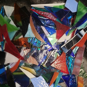 May include: A collection of colorful, irregularly shaped glass pieces. The fragments are in various shades of blue, green, red, orange, and white. Some pieces have a reflective, metallic appearance, while others are translucent. The pieces are scattered.